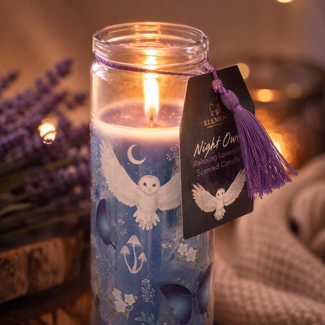 Candle in a glass jar with owl design and a tag, surrounded by lavender flowers.
